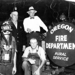 Four men. One in a gas mas. Next to Oregon Fire Department Rural Service sign